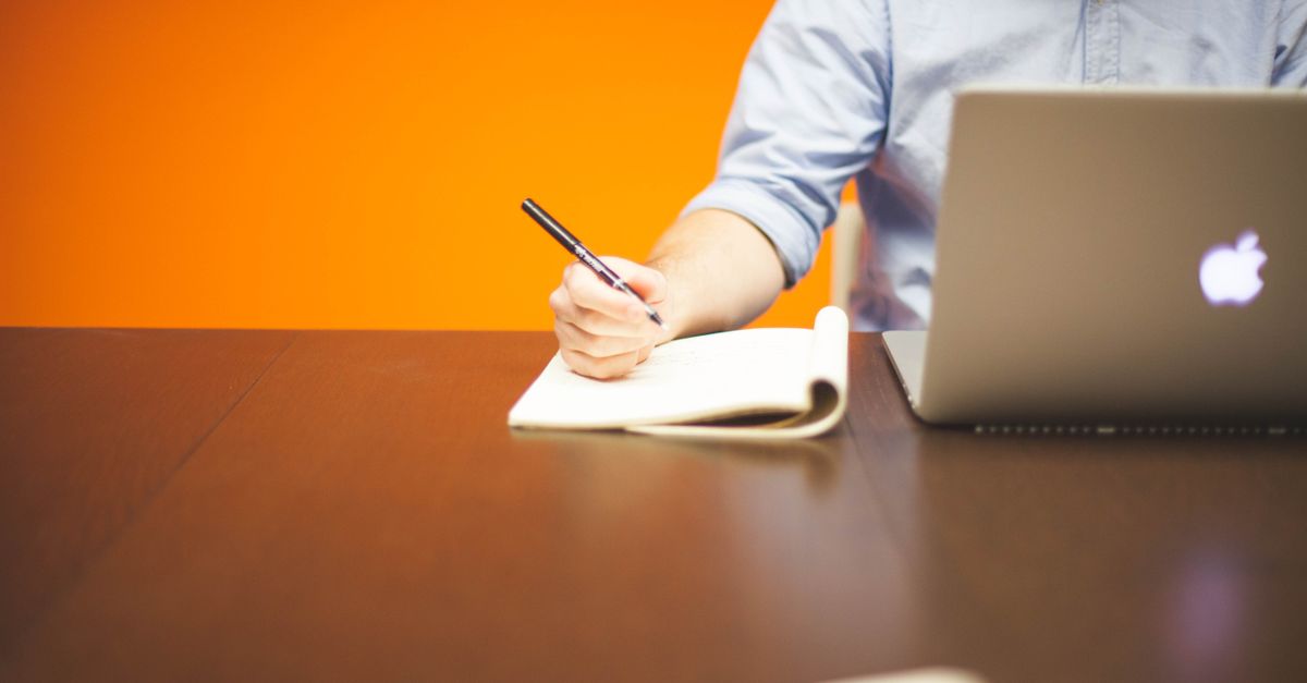 A person writing notes in a notebook beside a laptop on a wooden desk.