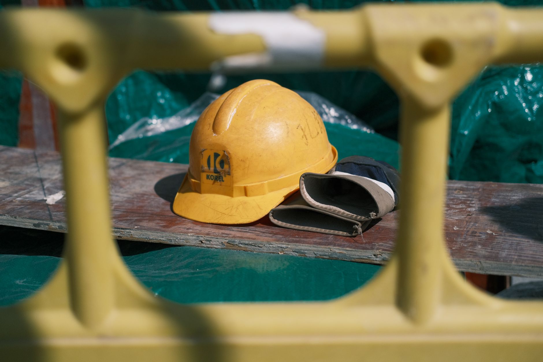 A yellow hard hat and work gloves resting on a plank at a construction site, seen through scaffolding.